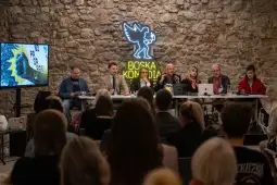 Panel dyskusyjny z uczestnikami przy stole, w tle kamienna ściana i neonowe logo.