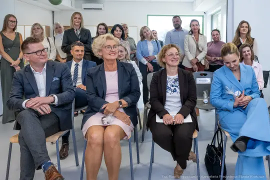 Grupa ludzi siedzących na spotkaniu, uśmiechnięte twarze, różnorodne stroje.