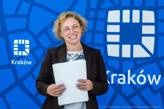Kobieta z dokumentami, uśmiechająca się na tle niebieskiej planszy z logo Krakowa.