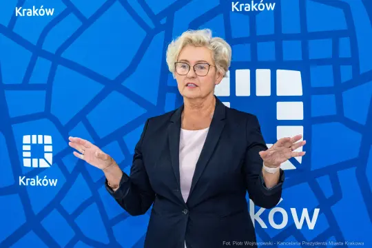 Kobieta w okularach w garniturze, gestykulująca na tle niebieskiego tła z logo Krakowa.