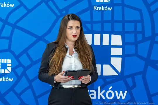 Kobieta w garniturze trzyma tablet, przemawia na tle niebieskiego tła z logo Krakowa.