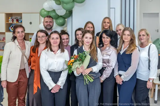 Grupa kobiet i mężczyzn w eleganckich strojach, z kwiatami i balonami w tle.