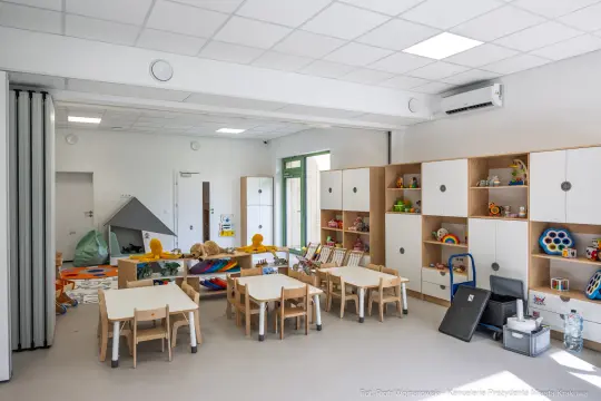 Nowoczesna sala przedszkolna z zabawkami, stolikami i regałami.