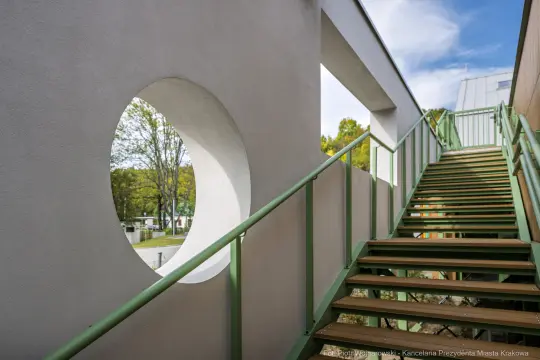 Schody prowadzące do okrągłego okna w nowoczesnym budynku. Zielone balustrady i jasne ściany.