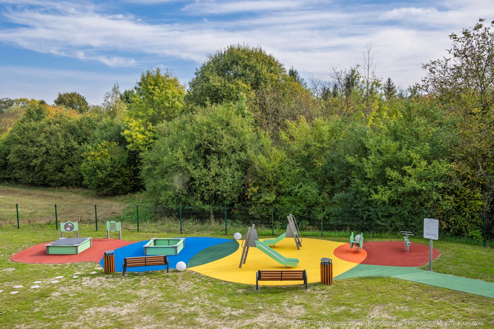 Plac zabaw z kolorowymi elementami, zjeżdżalnią i huśtawkami otoczony zielenią.  Autor: Piotr Wojnarowski