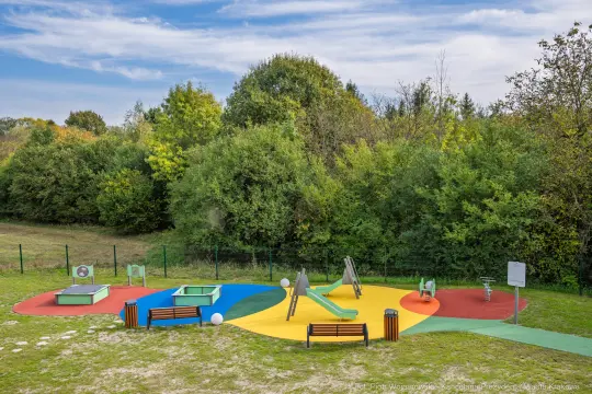 Plac zabaw z kolorowymi elementami, zjeżdżalnią i huśtawkami otoczony zielenią.