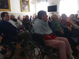Grupa seniorów w sali, część z nich na wózkach inwalidzkich, słuchająca wykładu.