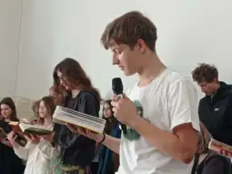 Młody mężczyzna czyta z książki, wokół niego grupa osób trzymających książki.