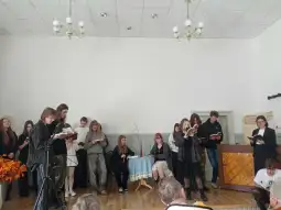 Grupa młodych ludzi śpiewa i recytuje teksty w sali z dekoracjami.