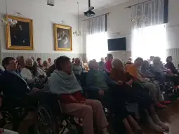 Grupa seniorów siedzących w sali, w tle obrazy i okna wpuszczające światło.