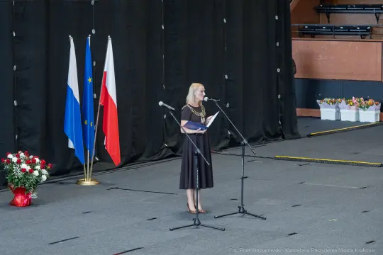 Kobieta w eleganckiej sukience przemawia na scenie, obok flagi Polski i Unii Europejskiej.
