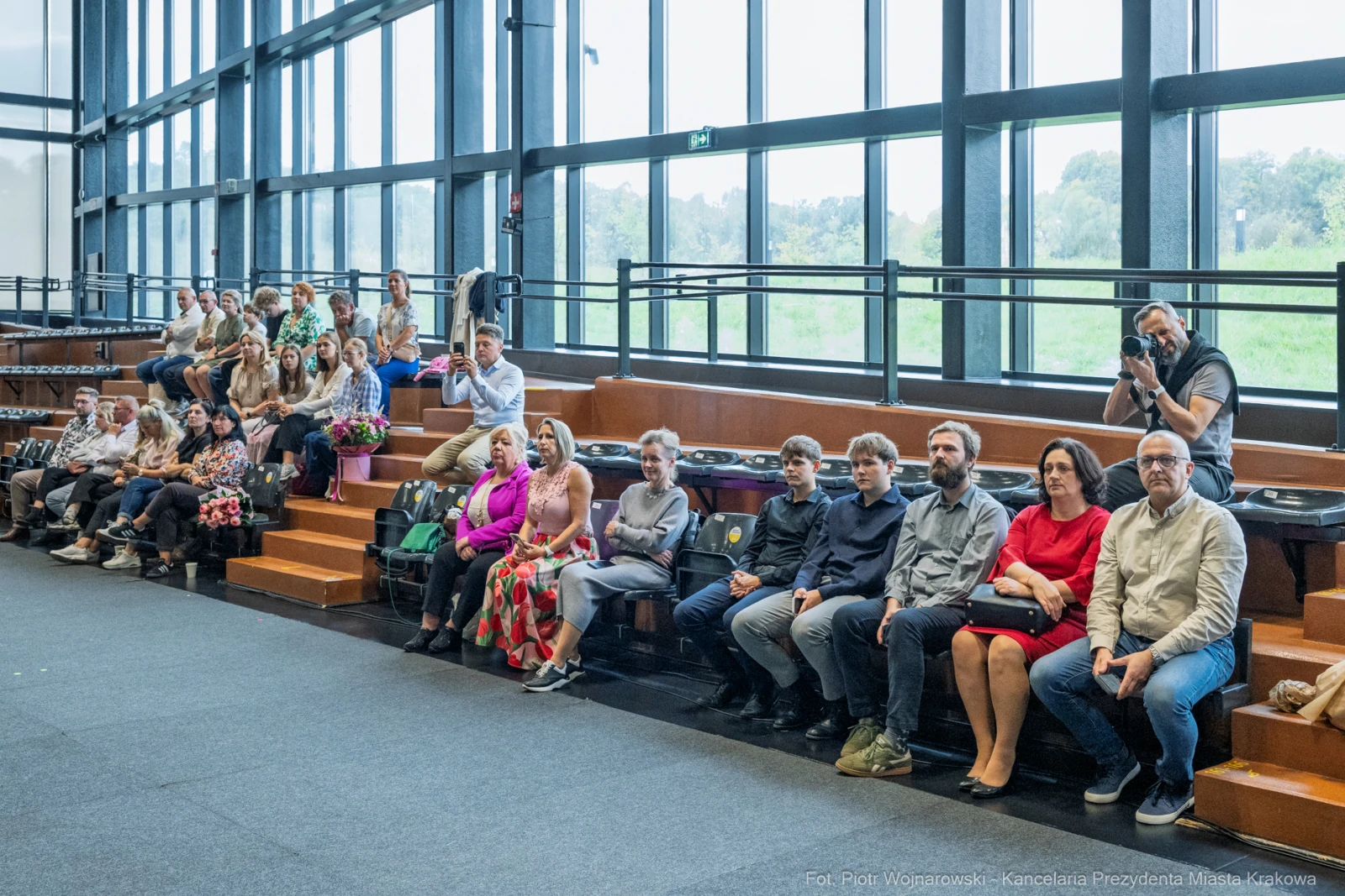 Grupa ludzi siedzących na schodach w sali, niektórzy trzymają kwiaty, inni rozmawiają.  Autor: Piotr Wojnarowski