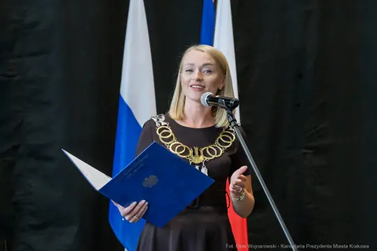 Kobieta w czarnej sukience z łańcuchem na szyi, przemawia do mikrofonu.