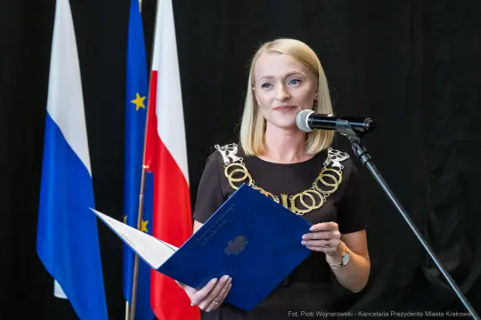 Kobieta w eleganckiej sukience z łańcuchem na szyi, przemawia do mikrofonu, trzyma dokument.