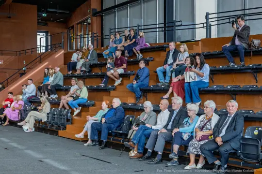 Grupa ludzi siedzących na widowni w sali, różnorodne reakcje i styl ubioru.
