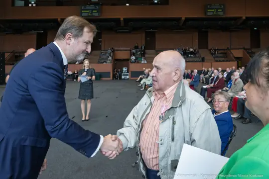 Spotkanie dwóch mężczyzn, jeden z nich podaje rękę drugiemu w sali z publicznością.