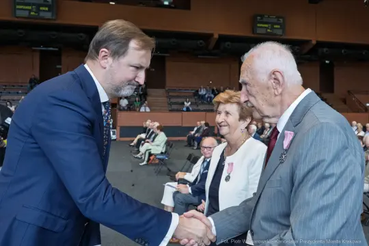 Uroczystość wręczenia odznaczeń, mężczyzna podaje rękę starszemu mężczyźnie, obok kobieta z medalem.