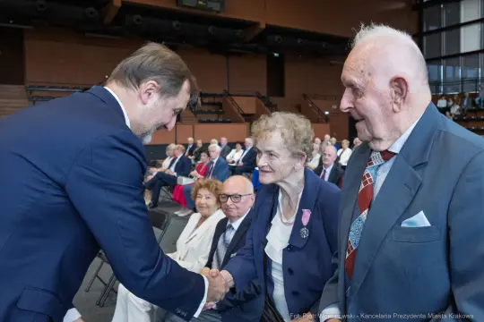 Uścisk dłoni między mężczyzną a starszą kobietą w towarzystwie innych osób.