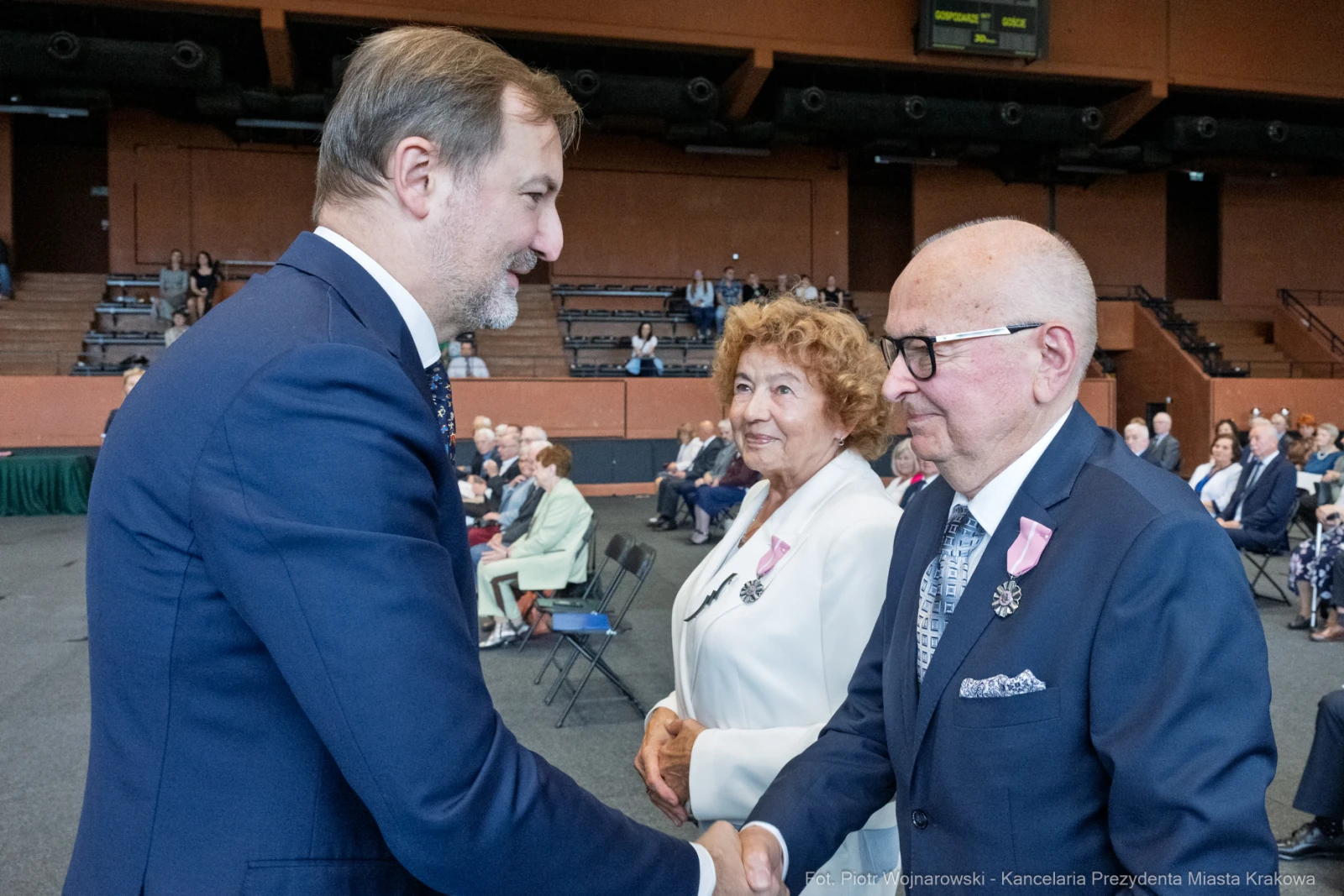 Uroczystość wręczenia odznaczeń, mężczyzna w garniturze wita starszego pana i kobietę.  Autor: Piotr Wojnarowski