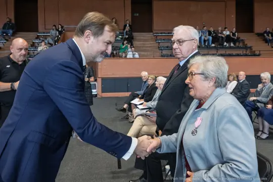 Uroczystość wręczenia odznaczeń, mężczyzna w garniturze podaje rękę starszej kobiecie.