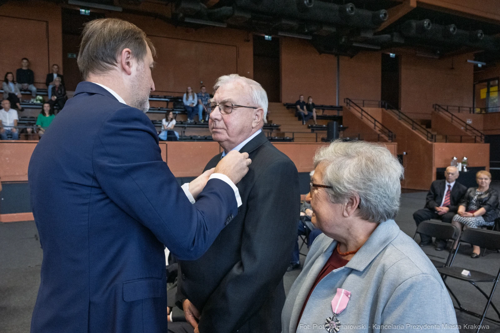 Uroczystość wręczenia odznaczeń, mężczyzna odbiera medal od drugiego mężczyzny, obok stoi kobieta.  Autor: Piotr Wojnarowski