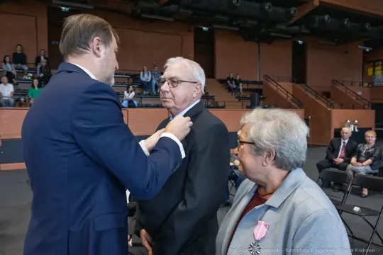 Uroczystość wręczenia odznaczeń, mężczyzna odbiera medal od drugiego mężczyzny, obok stoi kobieta.