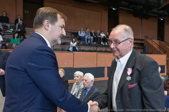 Mężczyzna w garniturze uścisnął dłoń starszemu mężczyźnie z odznaczeniem. W tle widownia.