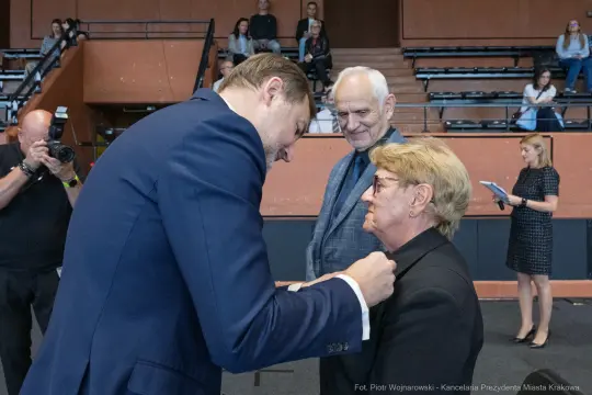 Wręczenie odznaczenia przez mężczyznę w garniturze starszej kobiecie. W tle widownia.