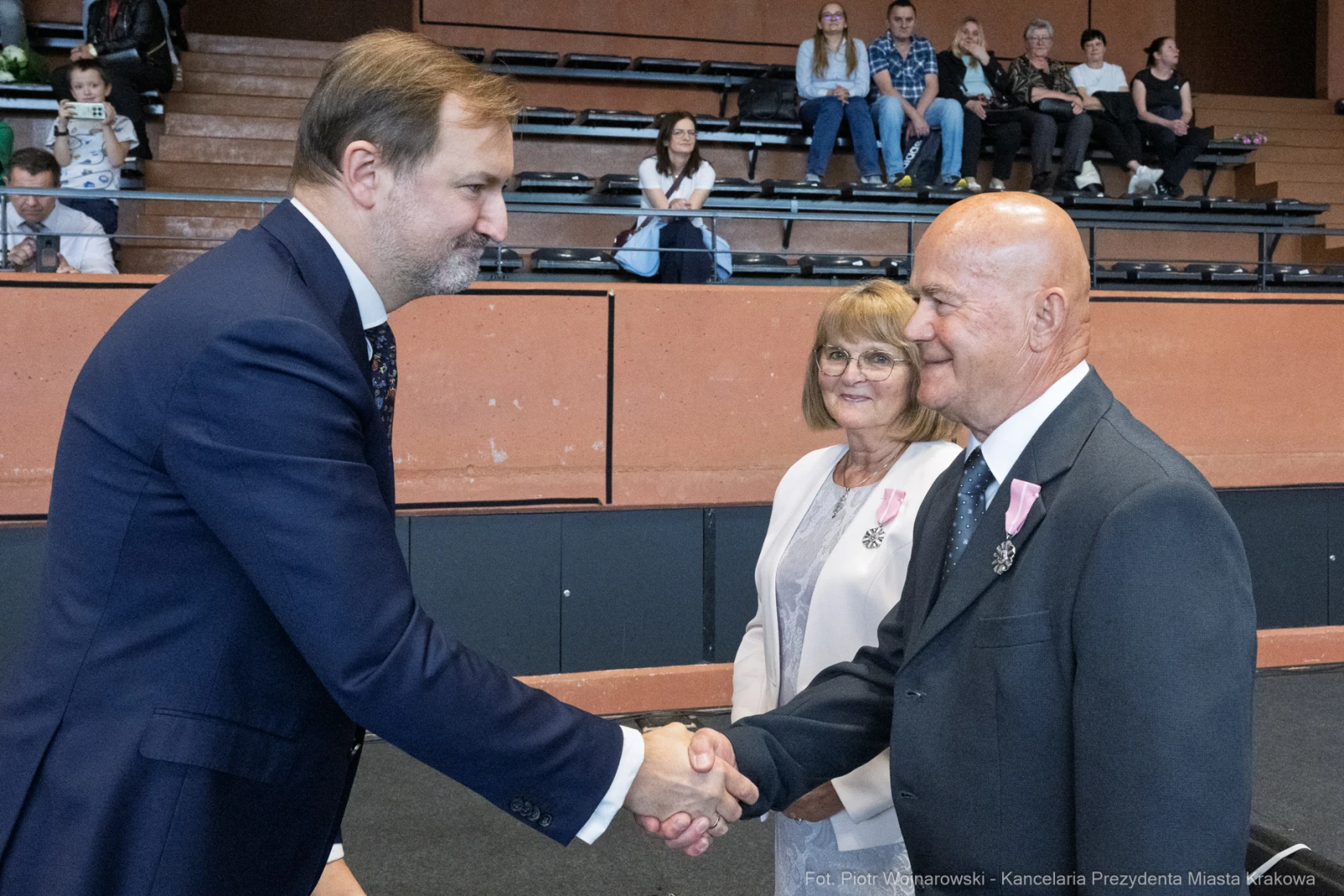 Mężczyzna w garniturze podaje rękę starszemu mężczyźnie, obok stoi uśmiechająca się kobieta.  Autor: Piotr Wojnarowski