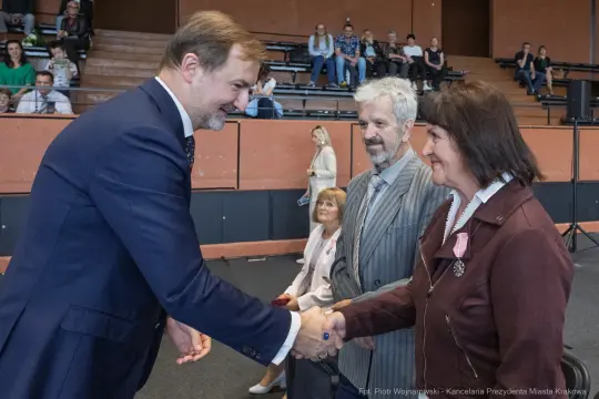 Mężczyzna w garniturze podaje rękę kobiecie, obok stoi starszy mężczyzna. W tle widownia.