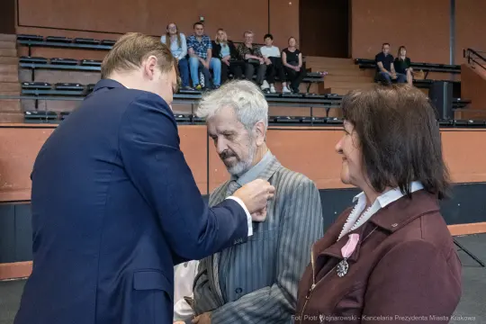 Ceremonia wręczenia odznaczeń w sali, dwie osoby w eleganckich strojach, mężczyzna odbierający medal.