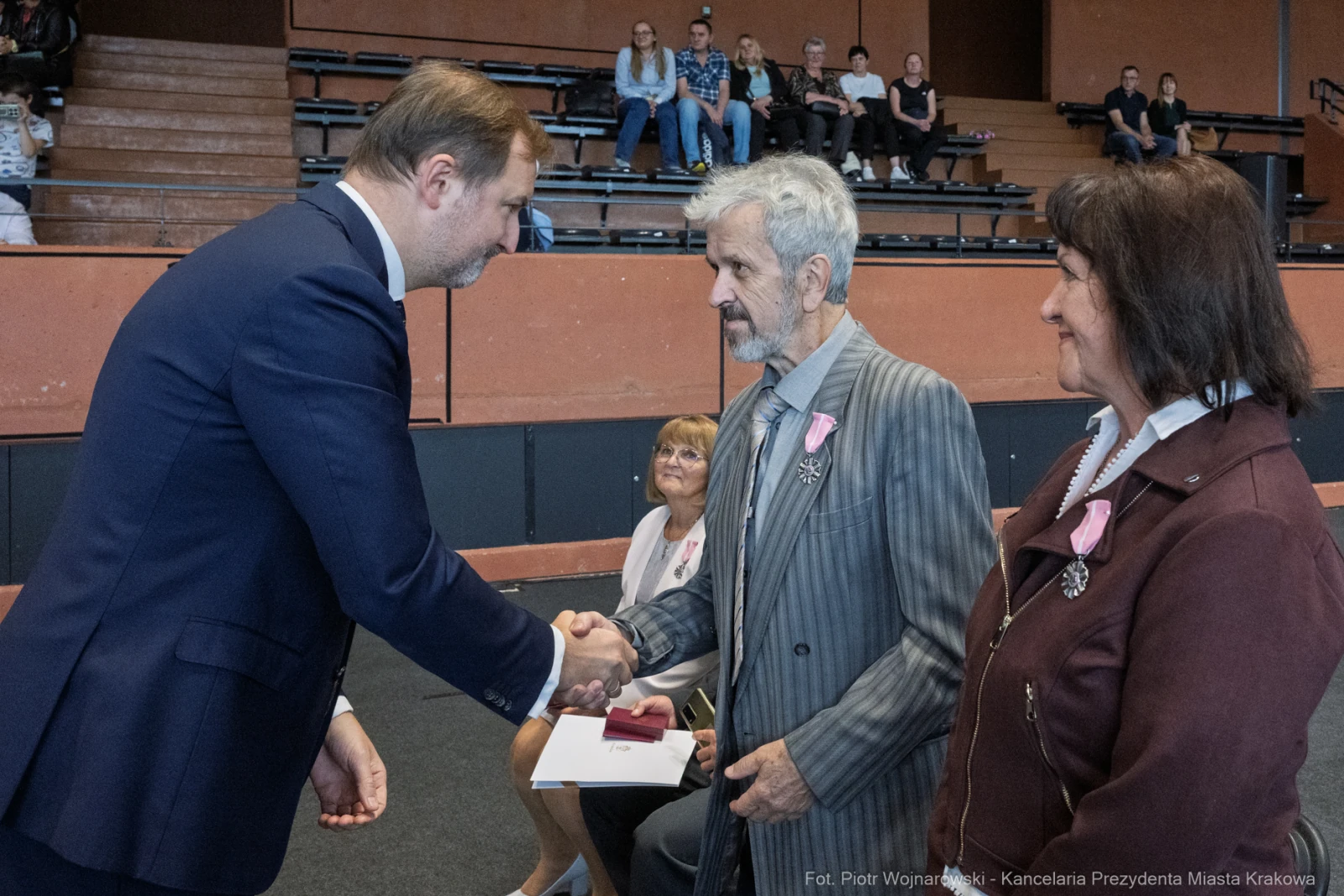 Uroczystość wręczenia odznaczeń, mężczyzna w garniturze podaje rękę starszemu mężczyźnie.  Autor: Piotr Wojnarowski