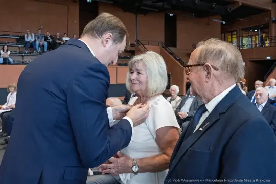 Wręczenie odznaczenia kobiecie przez mężczyznę w obecności starszego mężczyzny.