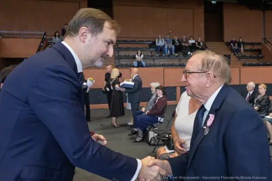 Uścisk dłoni dwóch mężczyzn podczas uroczystości w sali. W tle siedzą goście.