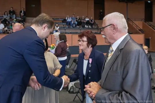 Uścisk dłoni między mężczyzną a kobietą w formalnej atmosferze. W tle widoczna publiczność.