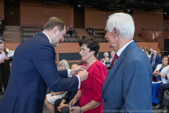 Wręczenie odznaczenia kobiecie w czerwonej sukience podczas uroczystości.