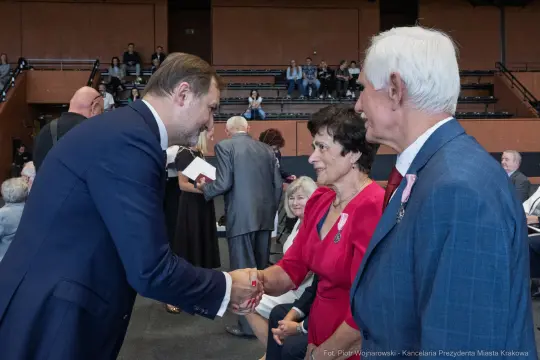 Uścisk dłoni między mężczyzną a kobietą w eleganckich strojach, w tle inni goście.
