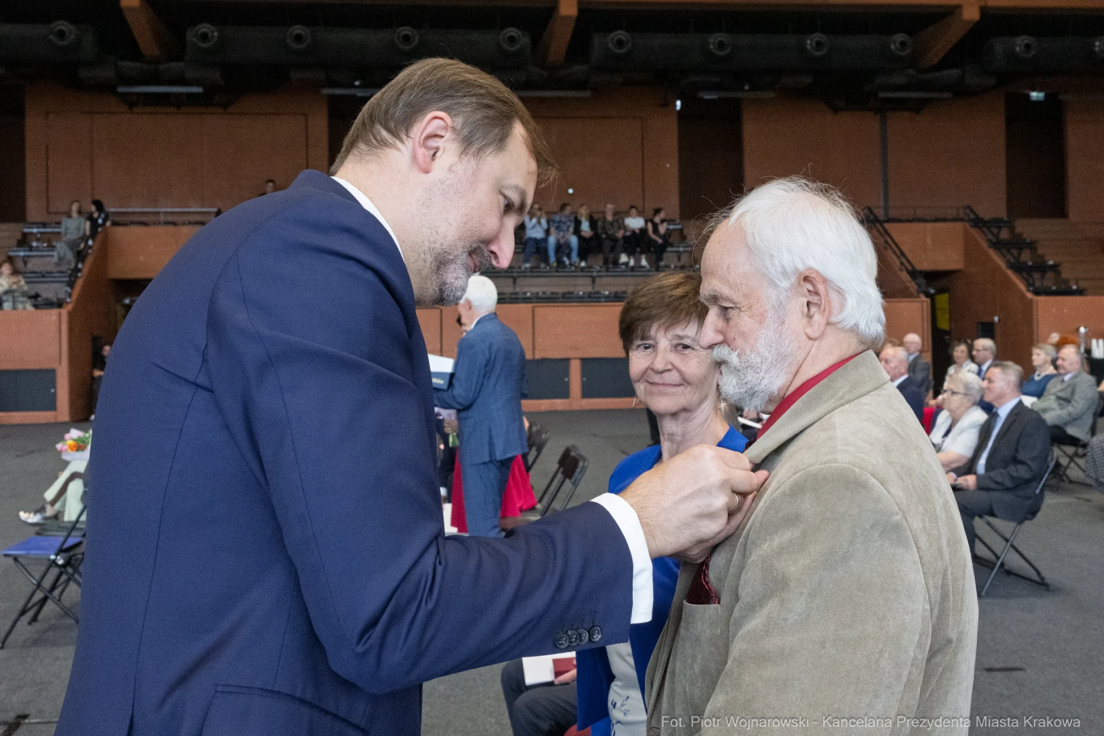 Wręczenie odznaczenia mężczyźnie przez innego mężczyznę, w tle uśmiechająca się kobieta.  Autor: Piotr Wojnarowski