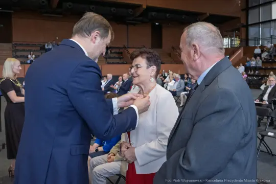 Uroczystość wręczenia odznaczenia kobiecie przez mężczyznę w obecności innych gości.