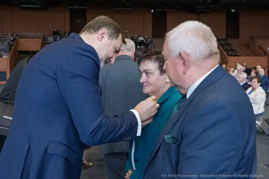 Mężczyzna przypina odznaczenie kobiecie, obok stoi starszy mężczyzna. W tle widoczna grupa ludzi.