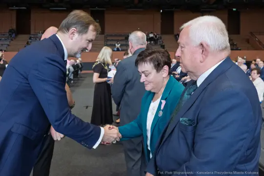 Mężczyzna w garniturze podaje rękę kobiecie w eleganckiej sukience, obok stoi starszy mężczyzna.