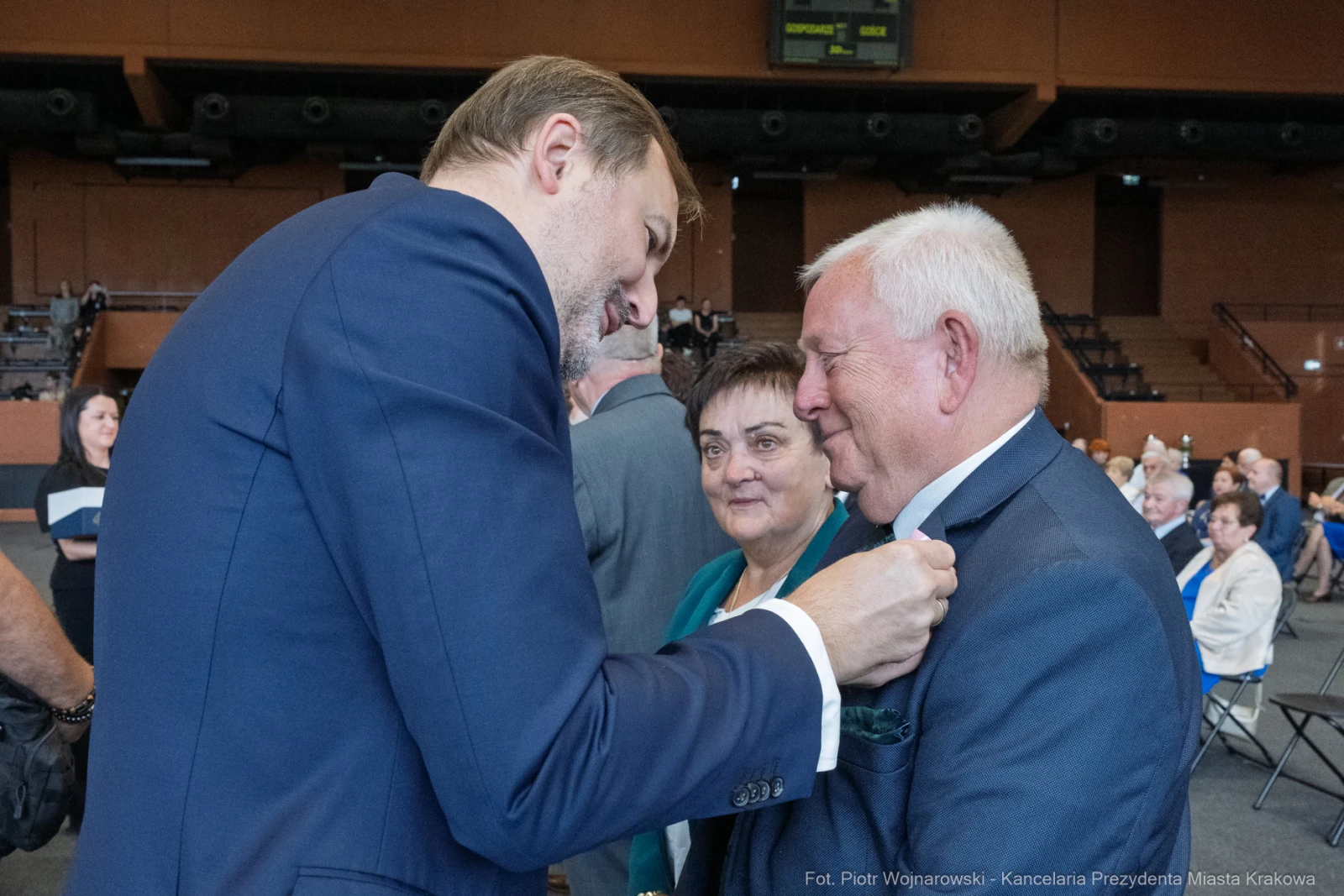 Mężczyzna przypina odznaczenie innemu mężczyźnie, w tle kobieta i inni uczestnicy wydarzenia.  Autor: Piotr Wojnarowski