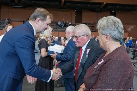 Mężczyzna w garniturze podaje rękę starszemu mężczyźnie w okularach, obok stoi kobieta.