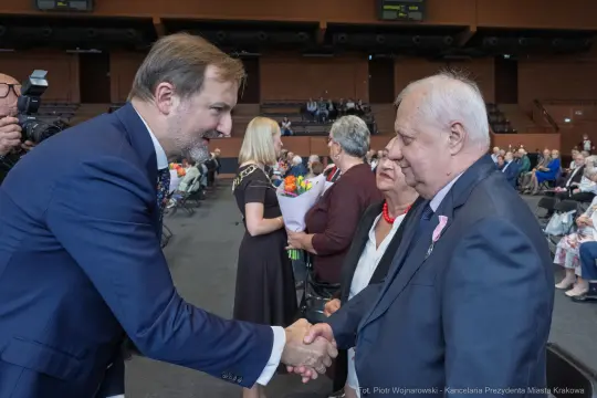 Mężczyzna w garniturze podaje rękę starszemu mężczyźnie w marynarce, w tle grupa osób.