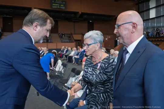 Spotkanie trzech osób w sali, uściski dłoni, widownia w tle.
