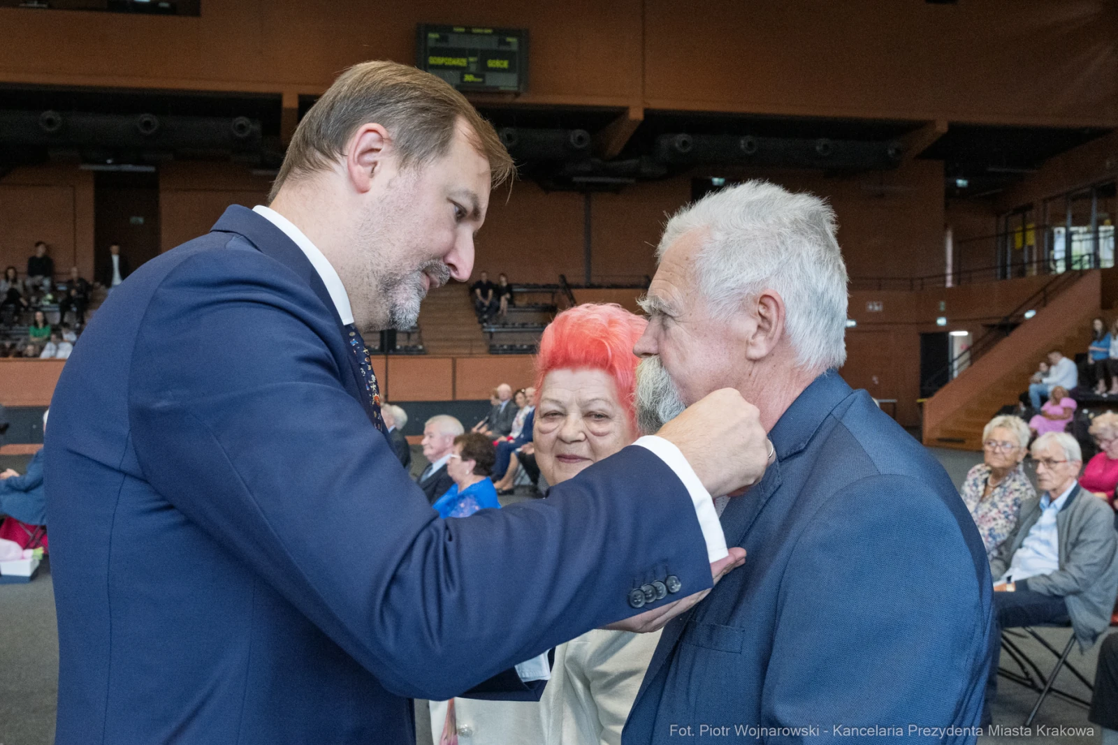 Mężczyzna przypina odznaczenie innemu mężczyźnie, obok stoi kobieta z czerwonymi włosami.  Autor: Piotr Wojnarowski