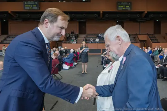 Uroczystość wręczenia odznaczeń, mężczyźni podają sobie ręce. W tle widoczna prowadząca wydarzenie.