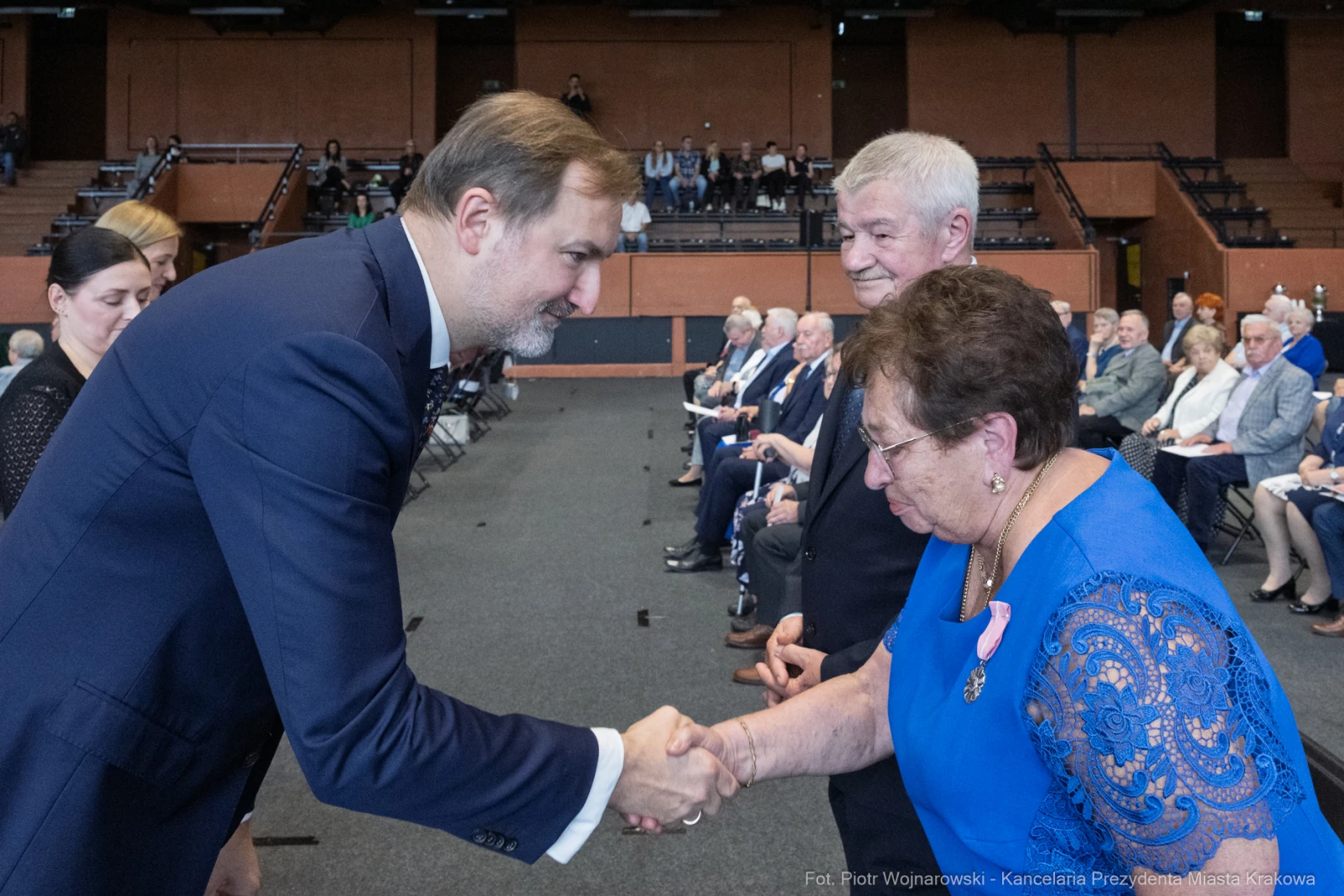Uścisk dłoni między mężczyzną a kobietą w niebieskiej sukience, w tle zgromadzenie ludzi.  Autor: Piotr Wojnarowski