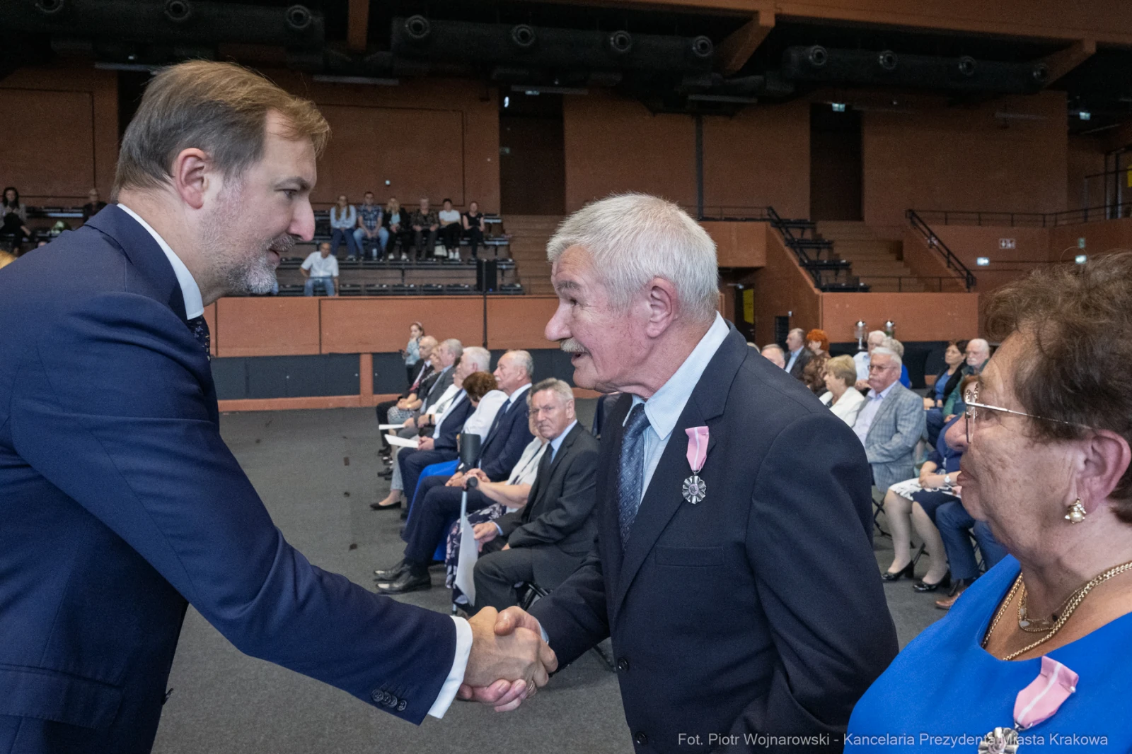Mężczyzna w garniturze podaje rękę starszemu mężczyźnie, w tle siedzą goście w formalnych strojach.  Autor: Piotr Wojnarowski