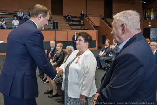 Uroczystość wręczenia odznaczeń, mężczyzna podaje rękę kobiecie w białej marynarce.